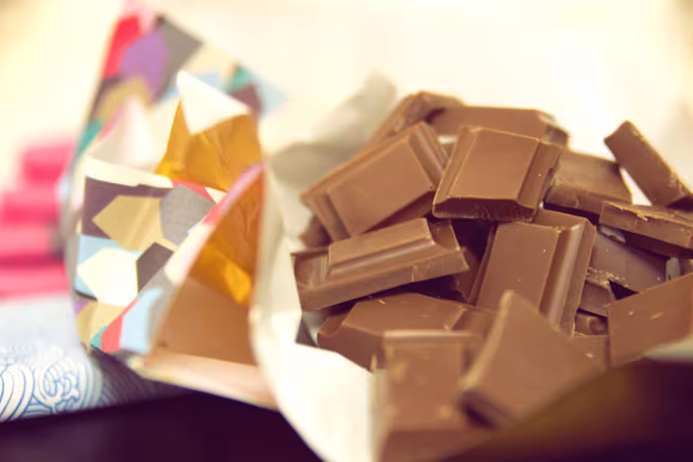 A close-up of several rectangular milk chocolate pieces piled together and spilling out of crumpled, colorful wrappers. The wrappers feature bright geometric patterns in shades of pink, yellow, blue, and red. The background is softly blurred with warm lighting.